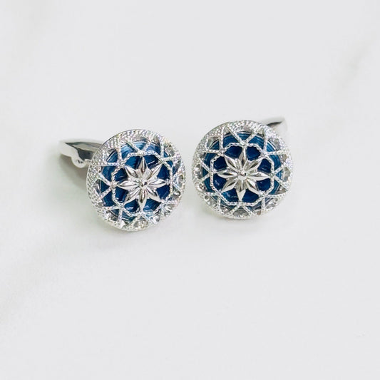 A pair of cufflinks with a silver floral filigree design over a blue background.