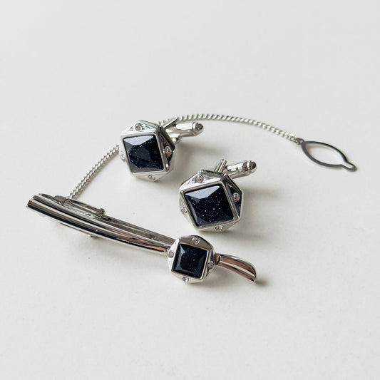 A set of silver-toned cufflinks and a tie clip with dark blue inlay and a chain link connector.