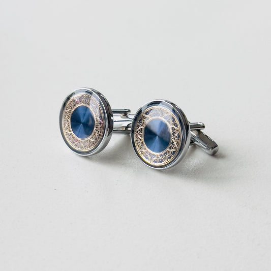 A pair of cufflinks with a blue cat's eye gemstone set in an ornate, silver and gold-toned circular frame with intricate designs.